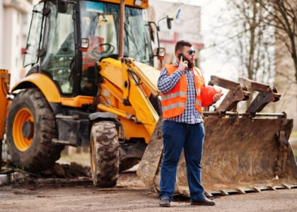 beard worker man suit construction worker safety orange helmet sunglasses against tractor with mobile phone hand